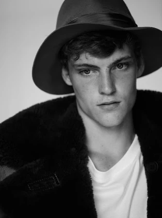 Close-up monochrome portrait of a young man wearing a wide-brimmed hat and dark coat over a white t-shirt against a plain background.
