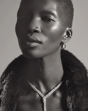 Close-up portrait of a Black woman wearing a diamond necklace and earrings with a dark textured collar against a grey background.