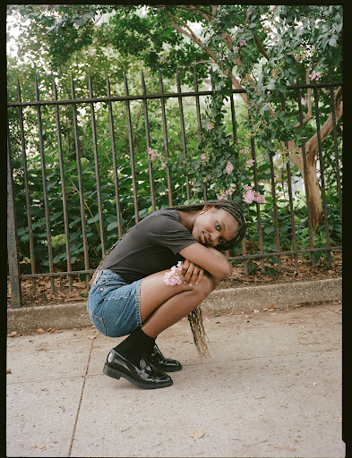 Woman squats on sidewalk in black shirt, denim shorts, black socks, and patent-leather loafers with a black metal fence in the background.