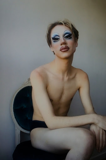 Person in drag makeup sits in ornate chair against a plain background, looking at the camera with a serious expression.