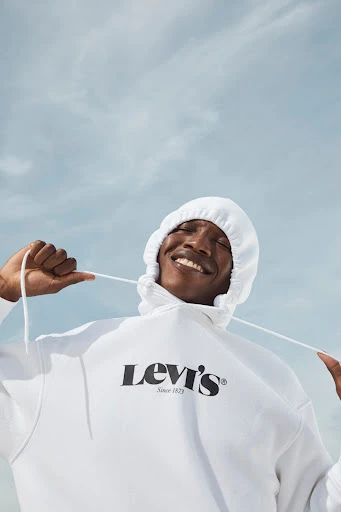 Smiling African-American man in white Levi's hoodie against a light blue sky backdrop, looking up with hands near the hoodie strings.