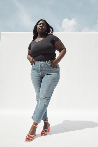 A fashionable plus-size African American woman stands indoors wearing a black top, jeans, and strappy pink heels against a white backdrop.