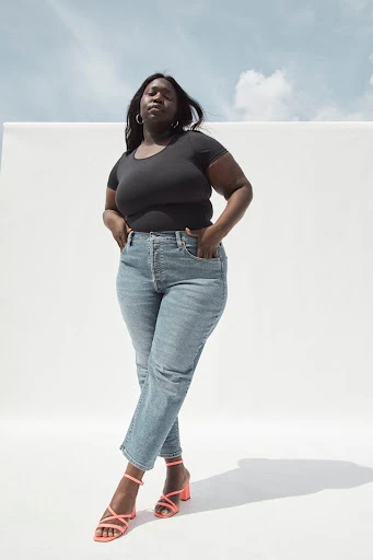 A fashionable plus-size African American woman stands indoors wearing a black top, jeans, and strappy pink heels against a white backdrop.