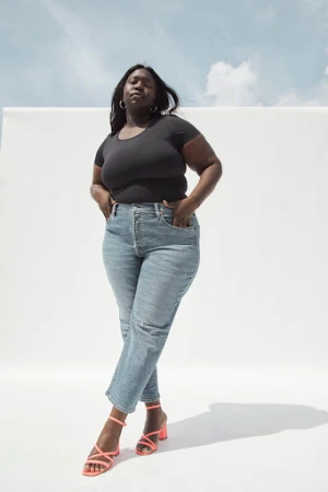 A fashionable plus-size African American woman stands indoors wearing a black top, jeans, and strappy pink heels against a white backdrop.