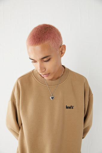 Headshot of a young adult with dyed pink hair wearing a tan Levi's crewneck sweatshirt and heart pendant against a white background.