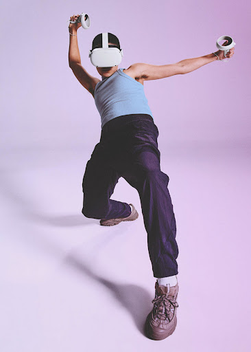 A person wearing a virtual reality headset with hand controllers poses in a studio, illuminated in bright pink and blue lighting.