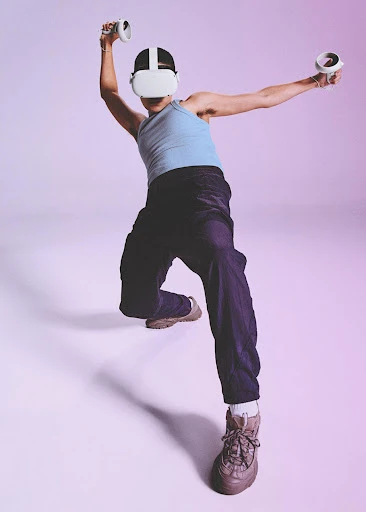 A person wearing a virtual reality headset with hand controllers poses in a studio, illuminated in bright pink and blue lighting.