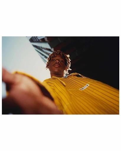 Low-angle shot of a young adult with amber-dyed hair wearing a yellow pinstripe shirt against a building and blue sky in an urban setting.