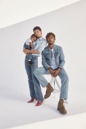 Family portrait in denim outfits: mother holding child, father seated on stool. White studio background, modern fashion, casual clothing.