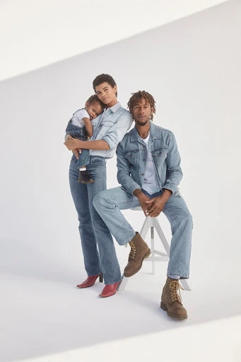 Family portrait in denim outfits: mother holding child, father seated on stool. White studio background, modern fashion, casual clothing.
