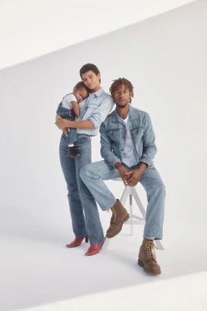 Family portrait in denim outfits: mother holding child, father seated on stool. White studio background, modern fashion, casual clothing.