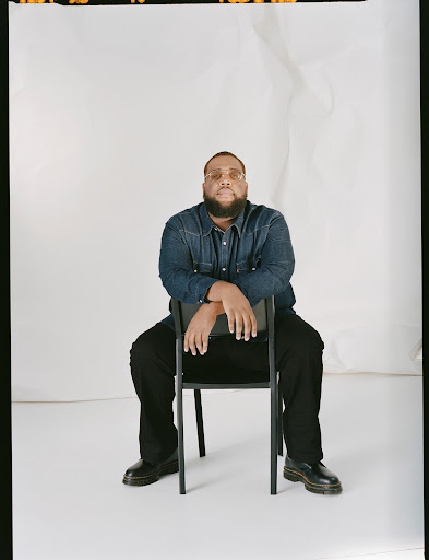 Portrait of a man sitting on a black chair in a photo studio, wearing a denim shirt, black pants, and shoes against a white backdrop.