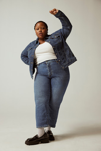 Full-length portrait of a plus-size Black female model in a denim jacket and jeans, standing with a raised fist against a plain backdrop.