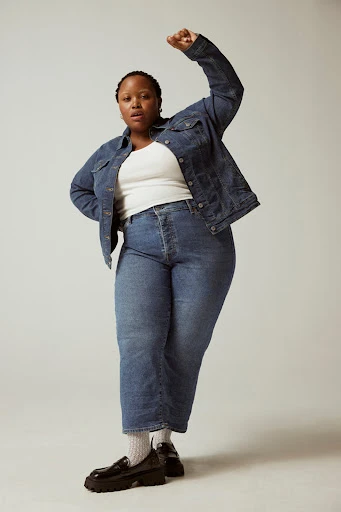 Full-length portrait of a plus-size Black female model in a denim jacket and jeans, standing with a raised fist against a plain backdrop.