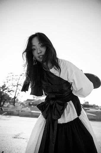 Portrait of a young Asian woman in a white dress with a black sash and skirt, looking at the camera against a bright sky background.