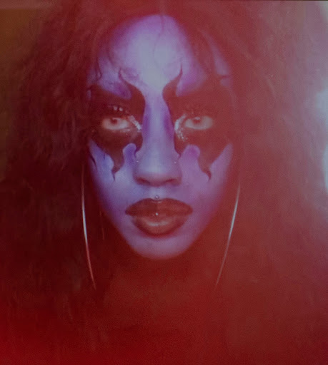 Close-up of a person with blue face paint and dramatic eye makeup, wearing hoop earrings, with dark hair, against a dark background.