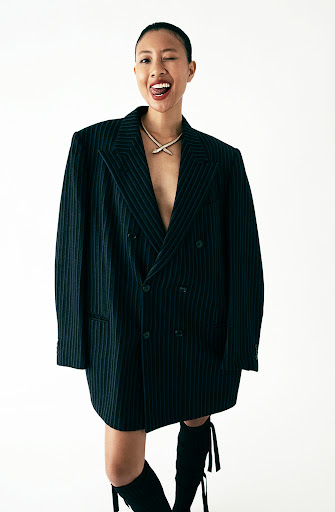 Adult woman in oversized pinstripe blazer and boots poses winking in a studio against a white backdrop with soft lighting.