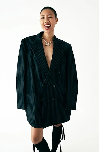 Adult woman in oversized pinstripe blazer and boots poses winking in a studio against a white backdrop with soft lighting.