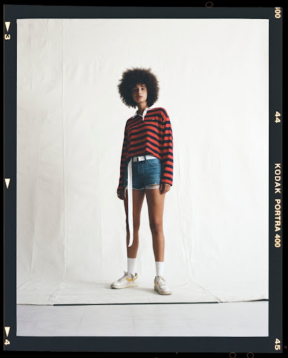 A young adult woman with natural dark hair stands in a photo studio wearing a striped rugby shirt, jean shorts, and sneakers against a white backdrop.