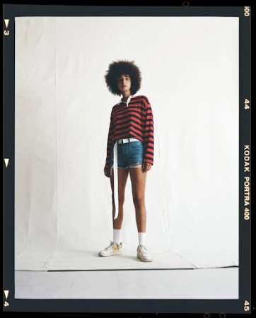A young adult woman with natural dark hair stands in a photo studio wearing a striped rugby shirt, jean shorts, and sneakers against a white backdrop.