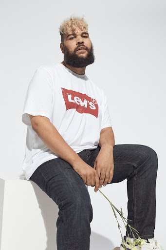 Plus-size male model with curly blond hair and beard wearing a white graphic t-shirt and dark jeans, holding flowers against a white background