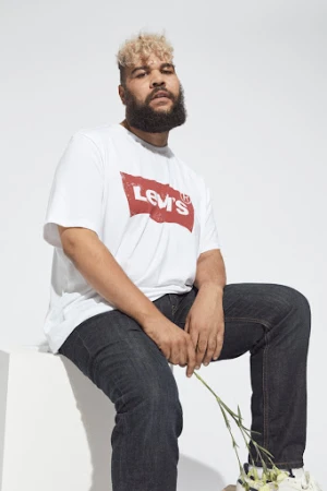 Plus-size male model with curly blond hair and beard wearing a white graphic t-shirt and dark jeans, holding flowers against a white background