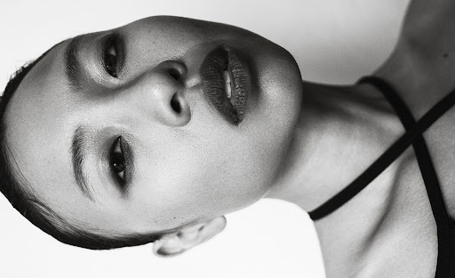 Monochrome portrait of a woman with dark hair and red lipstick, tilted perspective, black top, against a white background.