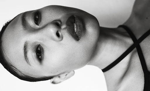 Monochrome portrait of a woman with dark hair and red lipstick, tilted perspective, black top, against a white background.