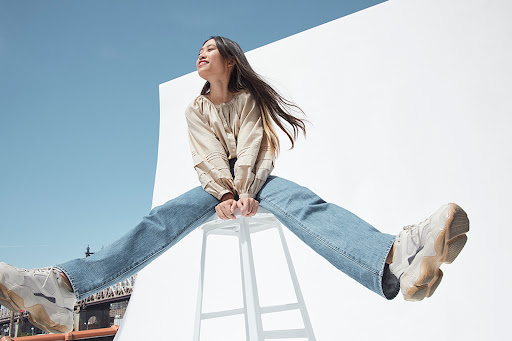 Low angle shot of an Asian woman in a beige blouse and blue jeans perched on a white stool against a bright sky background.