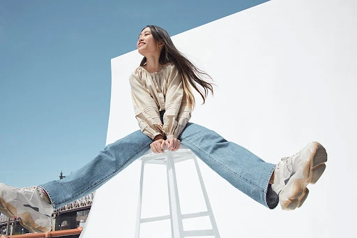 Low angle shot of an Asian woman in a beige blouse and blue jeans perched on a white stool against a bright sky background.