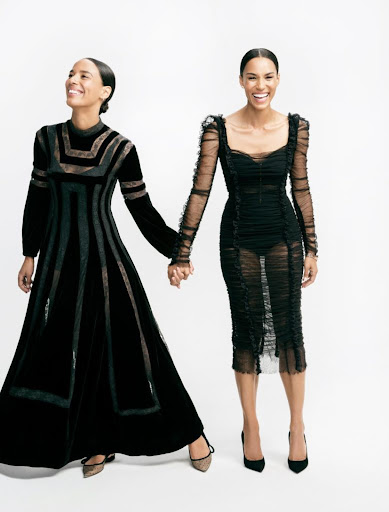 Two women holding hands in black formal dresses against a white background, smiling. Fashion editorial shot with elegant, dark-toned clothing.