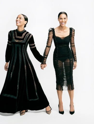 Two women holding hands in black formal dresses against a white background, smiling. Fashion editorial shot with elegant, dark-toned clothing.