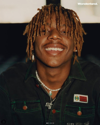 Close-up portrait of an adult male with dreadlocks smiling and wearing a green jacket and beaded necklace, with 'Wonderland' text visible.