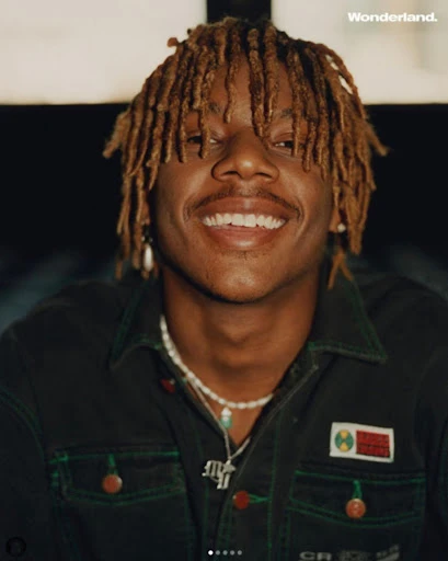 Close-up portrait of an adult male with dreadlocks smiling and wearing a green jacket and beaded necklace, with 'Wonderland' text visible.