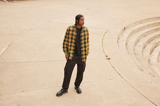 Man in plaid shirt and cargo pants stands on a concrete outdoor space, posing near stairs, looking to the side, with a neutral expression.