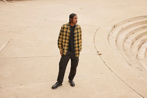 Man in plaid shirt and cargo pants stands on a concrete outdoor space, posing near stairs, looking to the side, with a neutral expression.