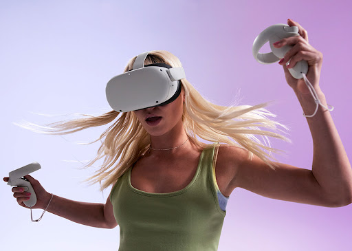 Woman with blonde hair is wearing a VR headset and holding controllers against a graduated purple background in a studio environment.