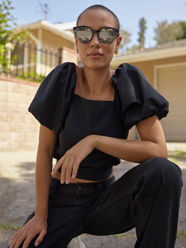 Woman with buzzcut and sunglasses posing outdoors in a black puff-sleeve blouse and black jeans kneeling in front of a house.
