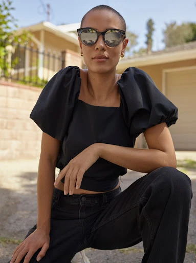 Woman with buzzcut and sunglasses posing outdoors in a black puff-sleeve blouse and black jeans kneeling in front of a house.