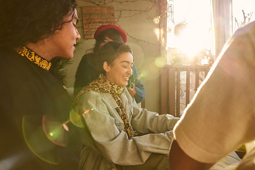 Group of friends gather in warm sunlight, laughing and talking; one woman wears a red hat, another a patterned scarf.