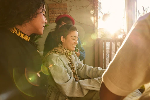 Group of friends gather in warm sunlight, laughing and talking; one woman wears a red hat, another a patterned scarf.