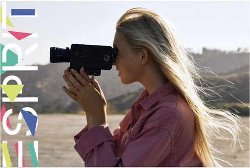 A woman with long blonde hair stands outdoors, looking through the viewfinder of an old-fashioned Super 8 movie camera. 