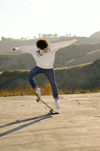 A young adult performs a skateboard trick outdoors with a hillside background on a sunny day, dressed in jeans and a graphic hoodie.
