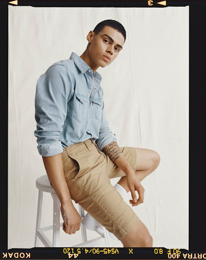 A young adult male model sits on a white stool wearing a light blue denim shirt and khaki shorts in a studio shot.