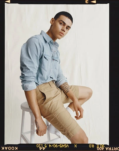 A young adult male model sits on a white stool wearing a light blue denim shirt and khaki shorts in a studio shot.