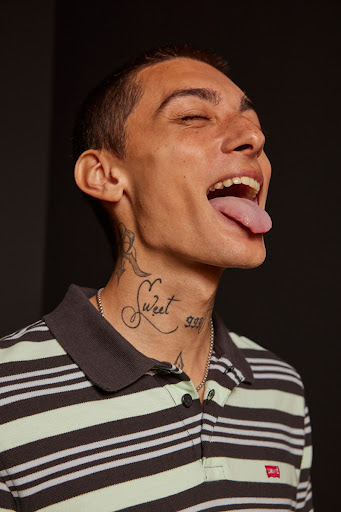 Portrait of a young adult male with a striped polo shirt, eyes closed, sticking out tongue, and neck tattoos against a dark background.