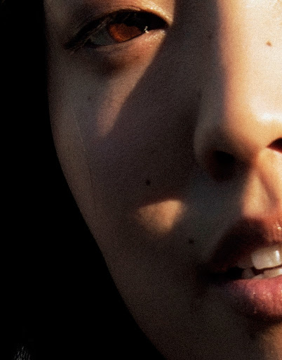 Close-up of an Asian female face with sunlight and shadow, showing an eye, nose, mouth, and fair skin with some moles. Dark background.