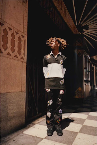 Young adult male in a vintage-inspired outfit poses on a city sidewalk near patterned masonry and dark doorway with radiating details.