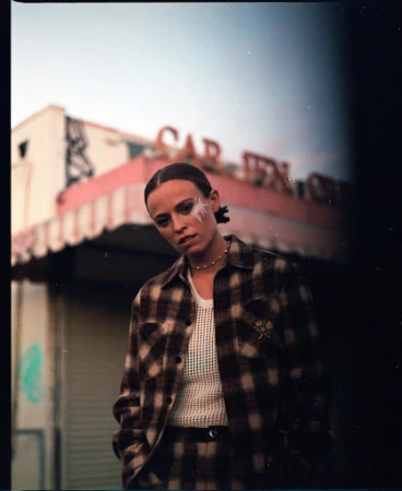 Woman in brown plaid shirt and matching trousers with face art standing outdoors in front of a building by Gena Tuso