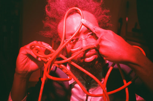 A man with an afro and glasses manipulates a red, coiled cable in a dimly lit indoor setting under bright, saturated red light.